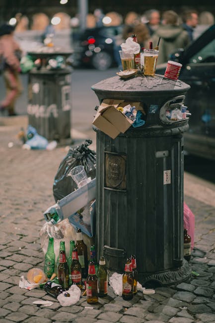 A black, cylindrical metal rubbish bin with a domed lid is positioned on a cobblestone pavement in an urban setting. The top of the bin is covered with various discarded items, including empty plastic cups with lids and straws, and a small cardboard box leaning against the side. Surrounding the base of the bin are several empty glass bottles, some still containing liquid, and scattered trash such as paper and plastic wrappers. In the background, there are blurred figures of pedestrians, parked cars, and additional litter, indicating a busy street scene. The environment appears to be a public outdoor area with typical urban waste accumulation from nearby activity, possibly from a nearby café or gathering spot. The scene is lit with natural daylight, highlighting the textures of the cobblestones, the metallic surface of the bin, and the various types of waste present, subtly connecting to the context of private or alternative rubbish collection methods evident in busy city environments.