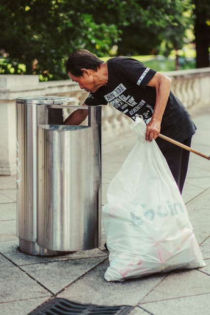 A middle-aged man with dark hair, wearing a black T-shirt and dark trousers, is seen carefully placing a large white garbage bag into a cylindrical stainless steel trash bin on a paved sidewalk. The trash bin has a shiny, reflective surface and is one of two identical bins positioned side by side in an outdoor setting. In the background, lush green trees and a low stone or concrete balustrade line the perimeter of the paved area, suggesting a park or public space. The man is holding a wooden stick or broom handle in his right hand, which rests against his hip. The lighting indicates a bright, sunny day, with natural daylight illuminating the scene. This outdoor rubbish collection activity reflects a focus on personal waste management and the use of private disposal methods outside of local authority collection points, aligning with the theme of independent rubbish removal services offered by companies like rubbishremovalelephantandcastle.com.