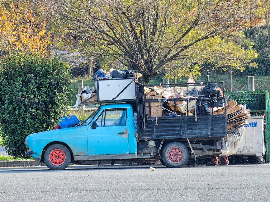 A small, light blue utility truck parked alongside a paved road with its cargo area fully loaded with various types of waste, including cardboard boxes, black plastic garbage bags, and miscellaneous debris. The truck's wheels are painted red, contrasting with the blue body, and the cargo is secured with ropes. Behind the truck, there is a large, leafy green bush and a tall tree with a broad canopy of green and yellow leaves, indicating a season of transition. The background includes a wire fence and a natural landscape with additional trees and foliage, suggesting an outdoor setting possibly near a park or residential area. The scene is illuminated by natural daylight, creating a clear and neutral atmosphere consistent with private waste collection or on-site rubbish clearing services often provided by independent rubbish removal companies like those at rubbishremovalelephantandcastle.com.