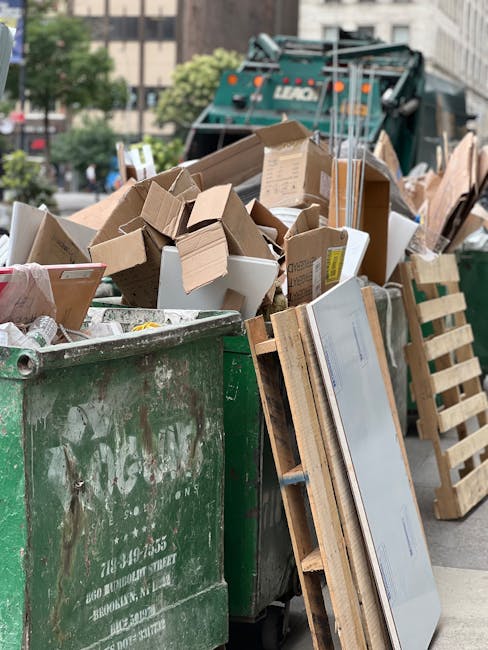 A large green commercial rubbish bin filled with a variety of waste materials, including scattered cardboard boxes in different sizes and shapes, some flattened and others still in their original form, along with a few white plastic containers. The cardboard exhibits various textures, from smooth coated surfaces to rough, corrugated edges, with some boxes bearing printed labels or tape. Leaning against the bin is a wooden pallet with visible slats and supports, slightly weathered but intact. In the background, a green waste collection vehicle with branded markings is parked on an urban street lined with trees, buildings, and parked cars, indicating a commercial waste collection site or on-site clearance operation. The overall scene is lit by natural daylight, creating a neutral, professional atmosphere typical of waste management services, observable within a city environment. The image captures an aspect of independent rubbish collection, consistent with private waste handling methods often used by local businesses or shops in the area.