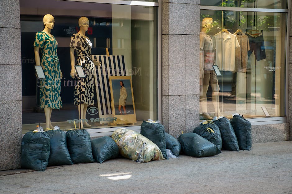 A row of dark green trash bags filled with rubbish is lined up on the pavement outside a retail store window in an urban setting. The bags are made of plastic, some appearing crumpled and others tightly knotted at the top, with a few revealing glimpses of mixed waste material inside. The store window behind the rubbish bags displays mannequins dressed in patterned dresses, along with framed promotional images and clothing racks visible through the glass. The building features a polished stone facade with a greyish finish, and sunlight casts soft shadows on the pavement. The scene suggests a temporary or spontaneous rubbish collection, likely part of private waste disposal services such as those offered by companies like rubbishremovalelephantandcastle.com, supporting local shop management or independent clearance efforts in the area.