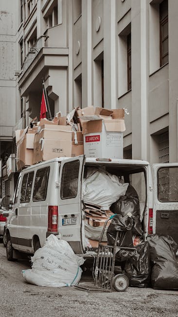 A white van parked on a city street beside a multi-story building with beige stone facades and rectangular windows. The van's rear doors are open, revealing an assortment of unmanaged waste including large black and white garbage bags, flattened cardboard boxes, and tangled packaging materials inside and just outside the vehicle. Several black garbage bags are seen leaning against the van's rear, some partially spilling onto the pavement. A metal trolley is positioned next to the open van, laden with additional rubbish. The scene suggests an on-site waste collection or private rubbish removal effort, as the load appears to be temporarily stored before disposal. The environment appears urban, with the building's architectural details visible in the background, and the lighting indicates natural daylight, typical of a busy city street providing a clear view of waste accumulation that may require professional rubbish removal services like those provided by [COMPANY_NAME].