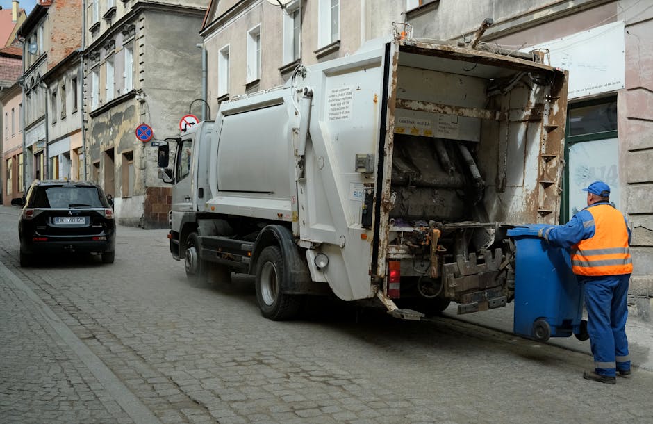A white commercial rubbish collection vehicle with a rusted metal rear loading compartment is parked on a narrow, cobblestone urban street adjacent to a row of older multi-storey buildings. The back hatch of the vehicle is open, revealing an empty, worn interior with visible rust and debris, indicating recent or ongoing waste collection. A worker dressed in a blue uniform, high-visibility orange vest, and blue cap is positioned to the right of the vehicle, actively pushing a blue wheeled bin towards the truck’s rear opening. In the background, a black car is parked parallel to the curb, and road signs indicating no parking or stopping are mounted on the building wall. The scene is lit by natural daylight, and the environment suggests an area where private waste collection services are provided, consistent with alternative rubbish removal arrangements near residential or commercial premises within the Elephant and Castle district.