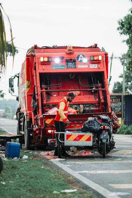 An individual in a high-visibility yellow and orange safety vest is operating a red rubbish collection truck on a street, with the rear loading mechanism open and debris visible underneath. The truck has a metallic finish with signs of use, and the back section features amber warning lights and labels indicating its registration number BRJ 7751 and operational details. The person appears to be clearing or loading waste, using tools and standing next to a small black scooter parked nearby, which has a black plastic bag and other items secured on its seat. The scene is set outdoors on a paved roadway with a grassy verge on the left, some trees, and utility poles in the background, under an overcast sky. The environment suggests an urban or suburban area, with elements consistent with private waste collection rather than municipal trash services, aligning with services offered by companies like rubbishremovalelephantandcastle.com for same-day rubbish removal and on-site clearance. The overall atmosphere is functional and orderly, with a focus on waste management activity.