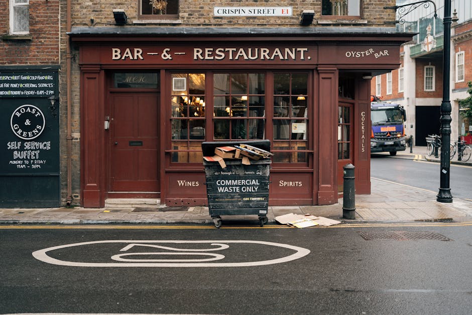 A black wheeled bin labeled 'Commercial Waste Only' is situated directly in front of the entrance to a red-brown wooden restaurant or bar, which features large multi-pane windows and signage indicating offerings such as wines and spirits. The bin appears to contain discarded cardboard boxes and paper, with some contents protruding and a few flattened boxes scattered on the pavement near its base. The setting is on a city street, with a double yellow parking restriction line visible on the road and a large white circular marking painted on the asphalt. To the left of the building, there's a black chalkboard sign advertising a self-service buffet, while to the right, a black bollard and a street lamp are positioned along the sidewalk. A delivery truck is partially seen in the background, and bicycles are parked nearby. The overall scene suggests an urban environment where commercial waste management, including private rubbish collection or on-site clearance, is relevant, and the presence of the waste bin highlights the importance of proper rubbish handling for local businesses.