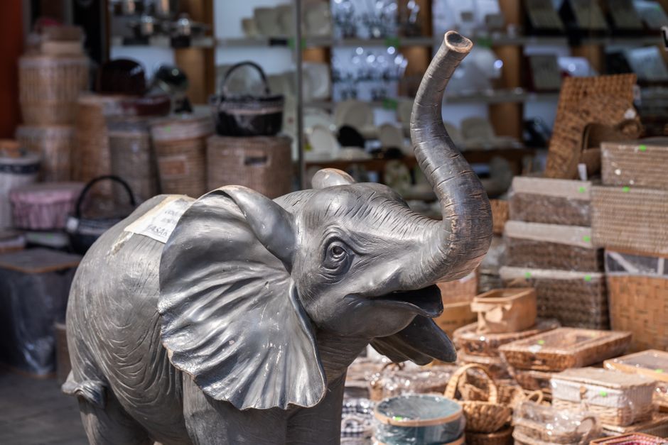 A life-sized, realistic sculpture of an elephant made from a dark grey, metallic-like material is displayed amid various wicker baskets and storage containers in a retail or warehouse setting. The elephant's surface features detailed textures that mimic the rough, wrinkled skin of a real animal, with prominent ears, a curved trunk raised upward, and expressive eyes. Surrounding the sculpture, there are stacked and neatly arranged wicker baskets of different sizes and shapes, some with lids and others open, showcasing their woven textures and natural light brown tones. In the background, shelves are lined with additional decorative items, including glassware and other household goods, suggesting a store or warehouse environment specializing in home or garden décor. The scene is well-lit with ambient lighting, highlighting the intricate details of the sculpture and the woven textures of the baskets. This setting appears to be an area dedicated to the sale or storage of decorative or functional household items, with occasional signs of potential on-site clearance or private disposal activities, which [COMPANY_NAME] may facilitate as part of their rubbish removal services in Southwark.